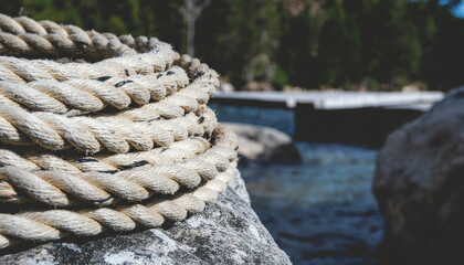 Fototapeta premium A heavy coiled mooring rope lies on weathered rocks beside a wooden dock, overlooking calm water and a misty pine shoreline, conveying a tranquil, rustic nautical atmosphere.