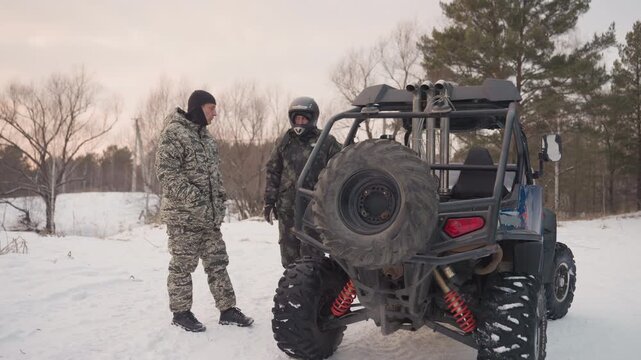 Snow atv duo converse beside utv in cold clearing, helmeted passenger inside and camo rider outside, route discussion and safety checks with spare tire visible, sunset glow and eager anticipation