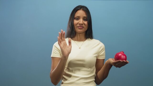 Woman holds red pomegranate in open hand and raises other hand palm out in studio; refusal restraint.