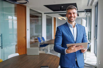 Fototapeta premium Professional businessman holding digital tablet, smiling in modern office, portraying success and leadership