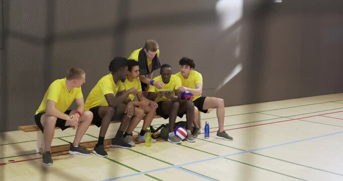 Diverse men in yellow tops sitting gym seat standing player talking, passing phone to show clip