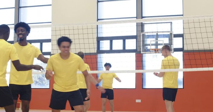 Diverse male team in yellow kit playing volleyball in gym, reacting to ball, regrouping to hydrate