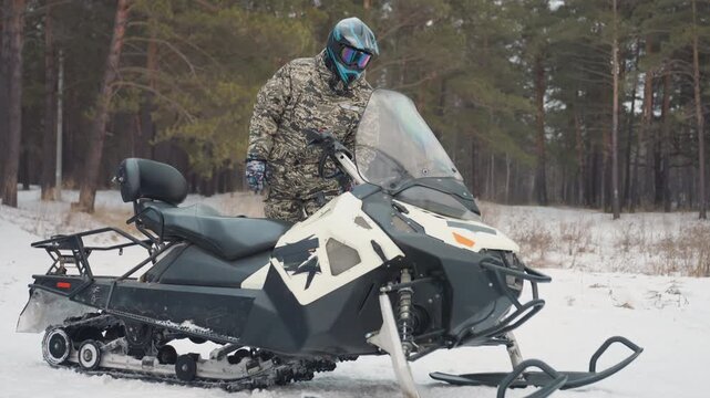 Snowmobile rider in snowy pine forest mounts machine and checks windshield, camo jacket with reflective visor, engine idling on frozen trail, winter overcast mood, tracks in powder, preparing