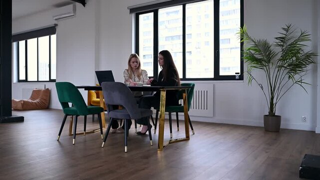 Two professional women sitting at a table and discussing business strategies, using a laptop and documents in a contemporary, sunlit office space with large windows and stylish furniture