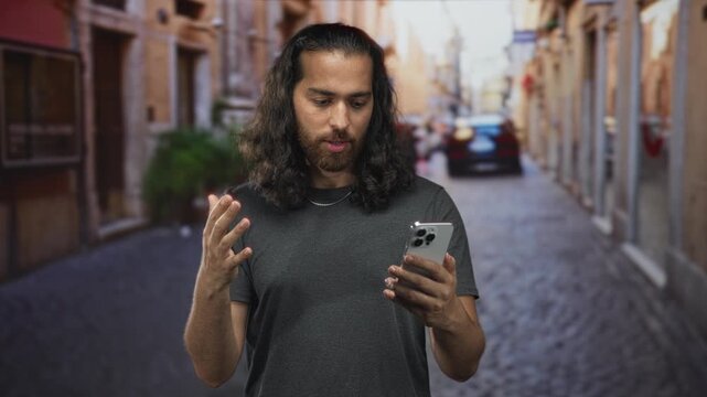 Man holding smartphone with raised hand and expressive gesture on a cobblestone street, looking at the screen and speaking; confusion troubleshooting.