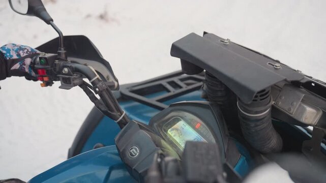 Atv handlebars and dashboard in snow, gloved hand twists throttle and turns ignition, instrument cluster lights up, rider prepares for offroad ride, front rack and mirror frame cold winter landscape,