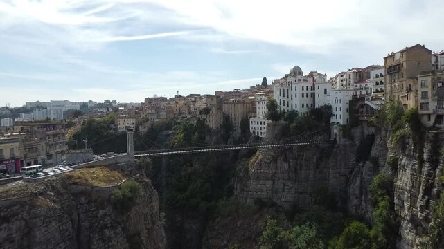 Ground-level aerial drone view of the Sidi M'Cid suspension bridge and the steep limestone cliffs of Constantine, Algeria, under a bright sky.