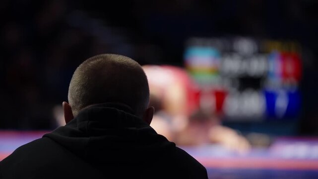 Silhouetted coach watches an intense wrestling bout as two athletes grapple on a brightly lit mat, their motions blurred in the background, capturing sideline focus and tension