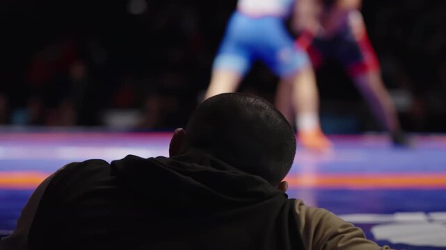 Focused coach watches intense wrestling bout from the sidelines as athletes grapple on a blue and orange mat under arena lights, capturing tense, strategic matside observation.