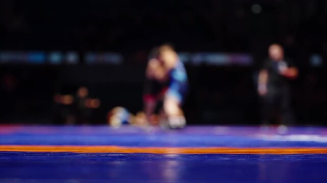 Low-angle view of a blue wrestling mat with an orange stripe as two blurred athletes grapple under arena lights, a referee nearby, capturing dynamic motion and intensity.