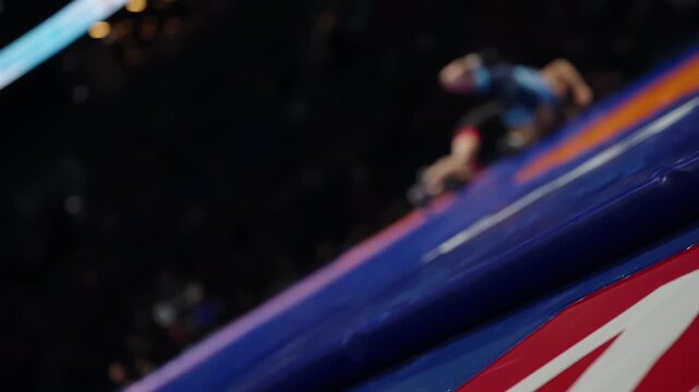 Low-angle view of a wrestling mat with vivid red and blue zones as two blurred athletes grapple in the background during an intense indoor bout, highlighting markings, padded edges, and dynamic motion