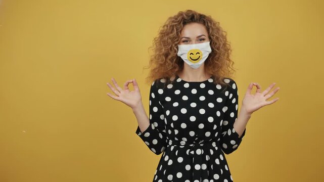 Female adult caucasian woman wears a protective face mask with a cheerful smiley emoji. She makes an okay gesture with both hands, expressing approval. Promoting health and safety