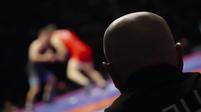 From a ringside vantage, a bald headed official observes an intense wrestling bout as two athletes in red and blue singlets grapple on a brightly lit mat inside a darkened arena.