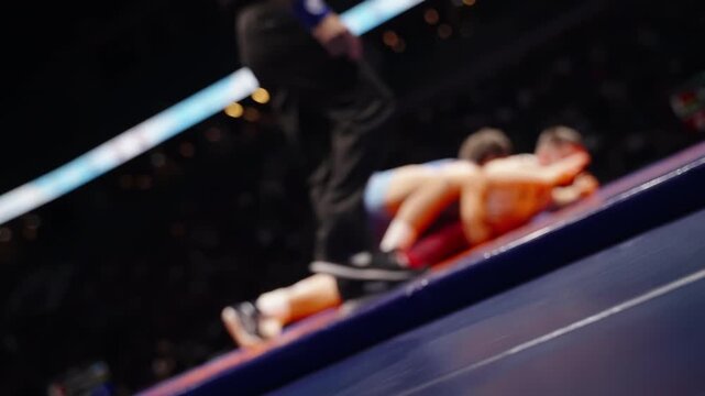 Defocused wrestling match on a brightly lit arena mat, as two athletes grapple and a referee steps in, capturing motion, intensity, and the competitive energy of a fast-paced bout.