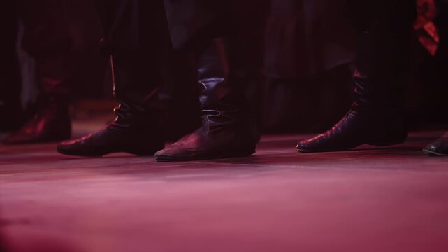 Low-angle close-up of synchronized dancers black leather boots striking a stage under dramatic red lighting, capturing rhythmic footwork and ensemble choreography in a dynamic theatrical folk