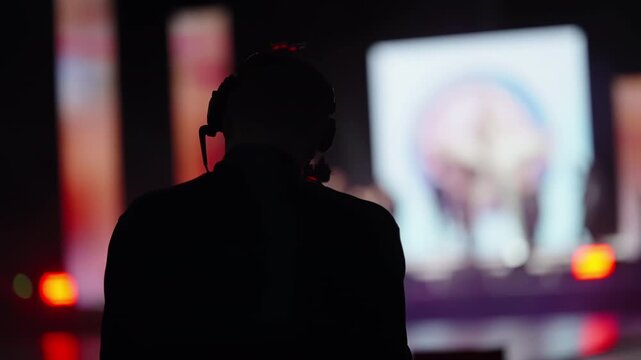 Silhouette of a headset-wearing stage technician overseeing illuminated screens and blurred performers during a live production, capturing backstage coordination and lighting
