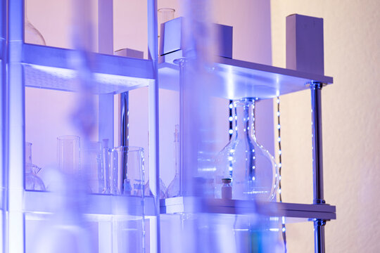 Chemical laboratory shelves with glass flasks and beakers under blue light, blurred foreground glassware, modern science workspace background with copy space.