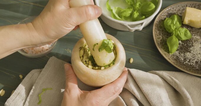 Hands grinding pine nuts and basil in marble mortar for Genovese pesto. Close-up of person using stone pestle to crush ingredients for traditional Italian sauce on wooden table. Making authentic pesto