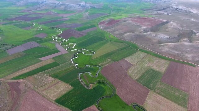 Aerial view of meandering river stream winding through green and brown farmland fields. Overhead footage shows sinuous creek channel curving across patchwork cropland on treeless floodplain.