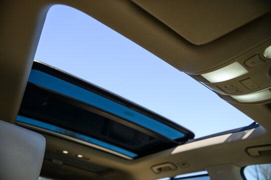 View of an open panoramic sunroof in a luxury car against a clear blue sky. Interior shot of automotive ceiling with glass roof and control panel in a modern premium vehicle
