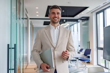 Fototapeta premium Confident businessman walking with laptop through modern office corridor, smiling and expressing success and professionalism
