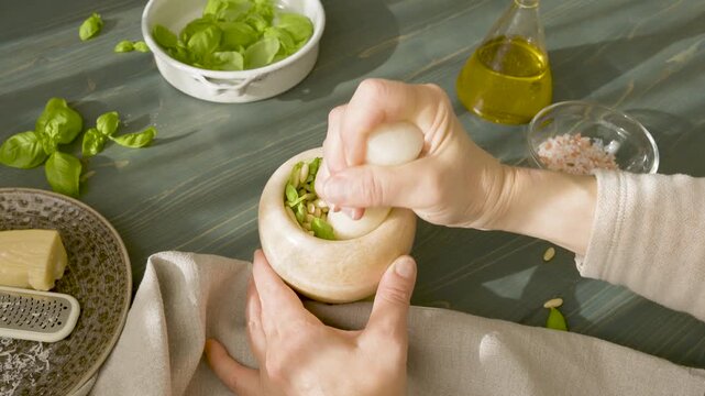 Hands grinding pine nuts and basil in marble mortar for Genovese pesto. Close-up of person using stone pestle to crush ingredients for traditional Italian sauce on wooden table. Making authentic pesto