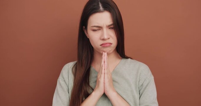 Sad young woman making a pleading gesture with hands together on a brown background.