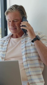 Vertical video: Phone ringing man talking, checking laptop by window, holding phone for work