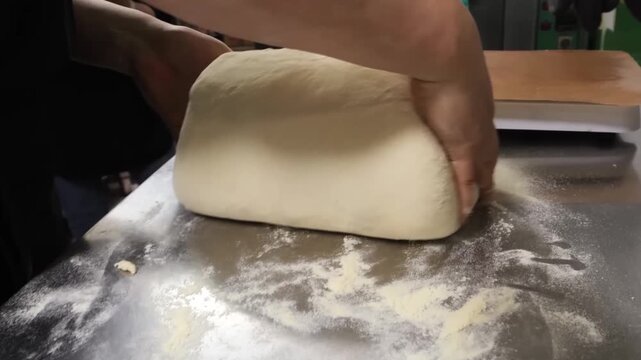 A close-up shot of a baker's hands kneading a large batch of fresh dough on a stainless steel counter dusted with flour.