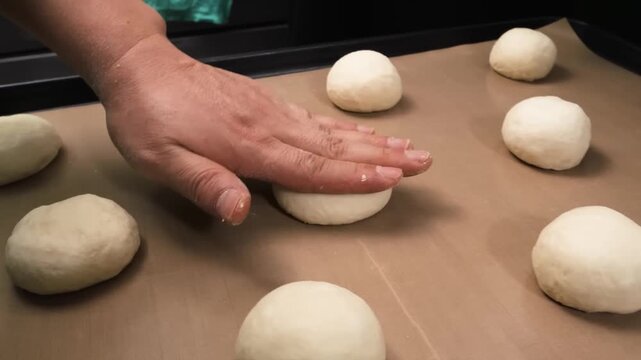 A close-up, high-angle shot of a person's hand flattening a ball of raw dough on a brown parchment-lined baking tray. Two other dough balls are visible, ready for preparation