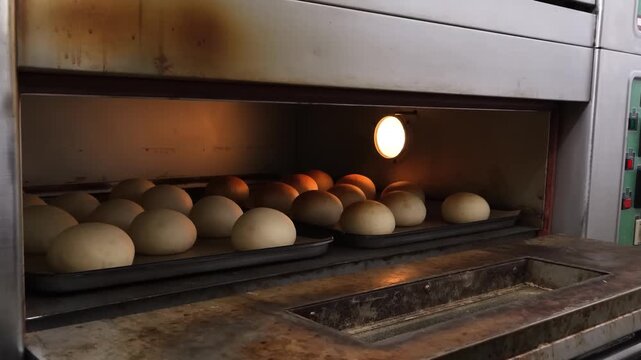 A close-up shot of multiple trays filled with dough rolls, baking inside a large, industrial-sized oven. The rolls are golden brown and rising, illuminated by the warm internal light of the oven.