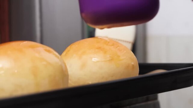 Close-up footage of a baker glazing freshly baked golden-brown buns or dinner rolls with a brush on a baking sheet. The warm, soft bread is ready for serving. Ideal for food, cooking, and baking-relat