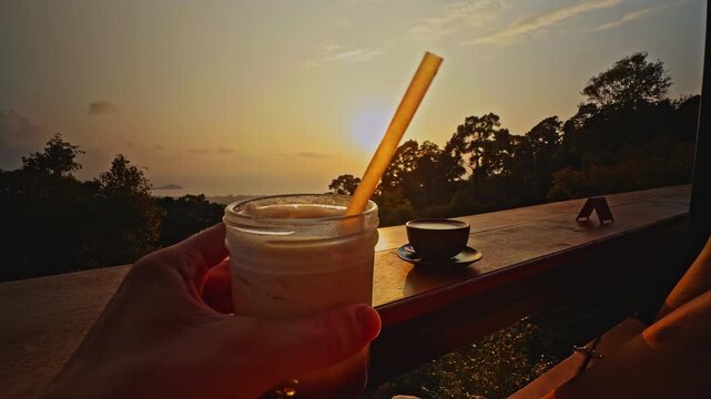 Hand Holding Iced Coffee Jar With Straw on Terrace at Tropical Sunset With Coffee Cup and Mountain Ocean Landscape Showing Relaxed Vacation Dining Experience