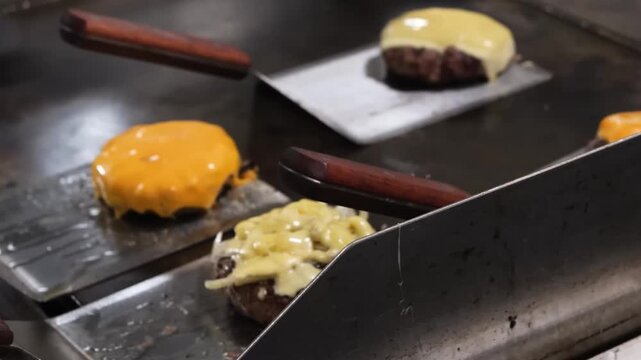 A close-up shot of a thick, gourmet beef burger patty cooking on a hot metal grill. A square slice of cheddar cheese is perfectly melted over the top, with visible steam and smoke rising as a spatula 