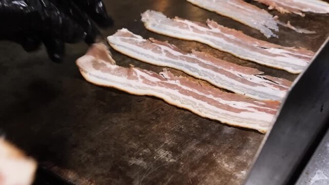 Close-up footage of a single strip of bacon sizzling and cooking on a flat, hot griddle or pan. The grease renders and bubbles around the meat, indicating high heat and the cooking process.