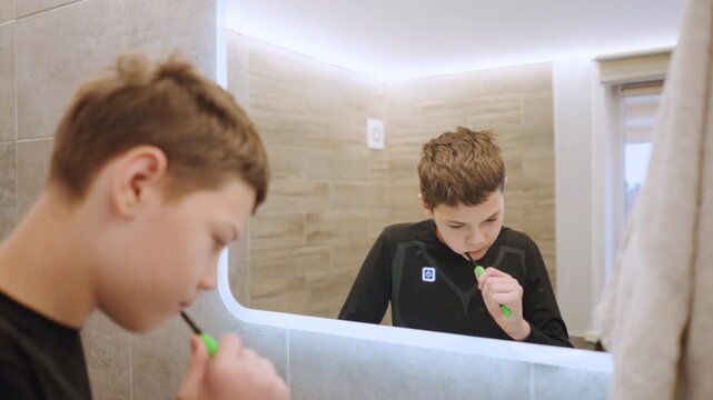 Young white grandson brushing teeth at mirror, quiet morning ritual in modern bathroom with LED backlit mirror, towel on rack, window light, toothpaste foam on brush, focused expression, domestic