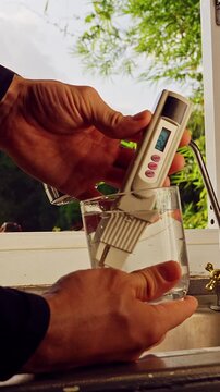 First Person View Of Hands Testing Drinking Water With Digital Quality Meter Showing Twenty Two Ppm In Glass Near Kitchen Faucet And Window With Tropical Vegetation Outside. Clean Drinking Water Verif
