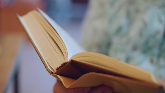 Close book pages in hands, elderly grandma reading quietly in cozy home, warm sunlight on textured pages, slow motion thumb flipping vintage paperback, intimate detail of binding and soft focus
