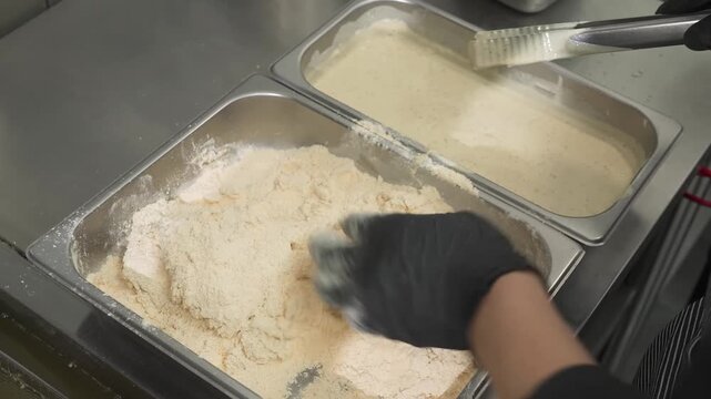 Close-up shot of a person wearing black gloves, using their hands to thoroughly coat a piece of raw chicken in a large metal container filled with a dry flour breading mixture. The setting is a profes