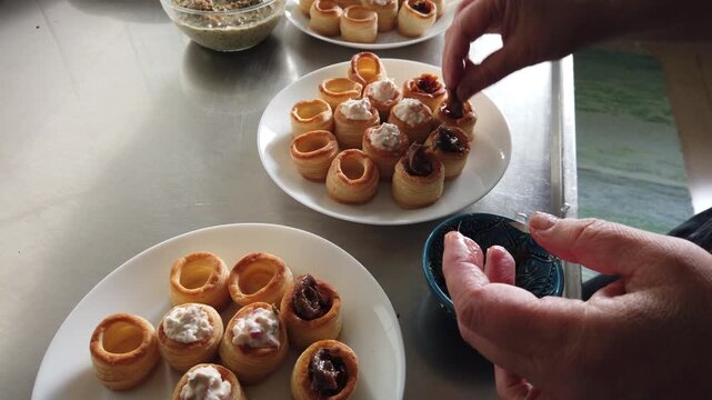 A hand arranges golden puff pastry shells filled with a creamy mixture on white plates beside a glass bowl in a stainless steel kitchen, evoking casual entertaining, hors d'oeuvres preparation and pla