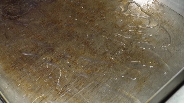 High-angle, close-up shot of cooking oil or liquid fat spreading and shimmering on the surface of a professional stainless steel griddle or baking sheet as it heats up.