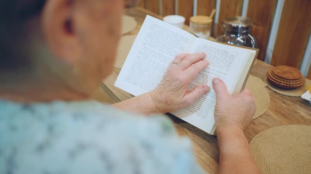 Closeup hands reading braille book at kitchen table with warm light, white grandma tracing raised dots with patient fingers, woven placemat, teapot and cookies nearby, calm focused atmosphere