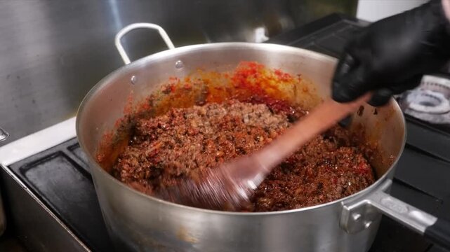 A close-up shot of a rich, homemade meat sauce (like bolognese or ragu) cooking in a stainless steel pot on a stove. The mixture of ground beef, onions, carrots, and a red sauce is being stirred with 