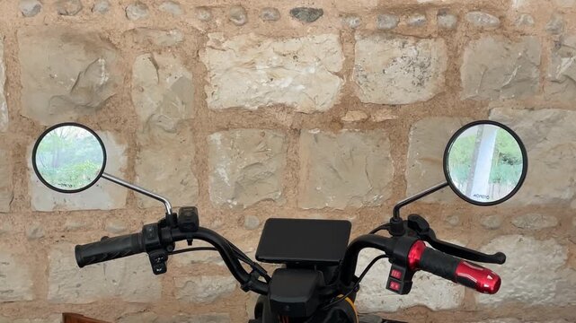 Motorcycle handlebars against a rustic stone wall background, featuring mirrors and controls
