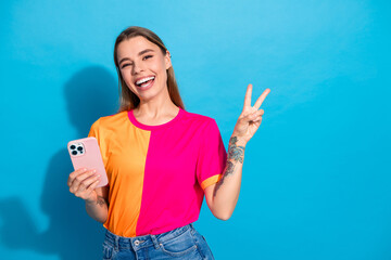 Young woman with colorful tshirt smiling and making a peace sign while holding a smartphone against a bright blue background for lifestyle advertising and shopping
