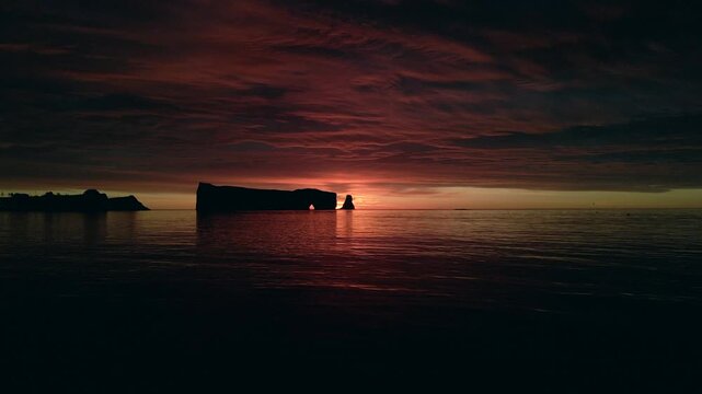 Cinematic aerial view of perce rock at sunset with dark red clouds reflecting in ocean
