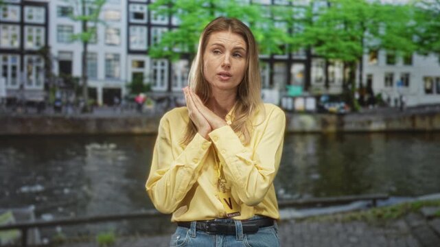 Woman wearing a visitor card lanyard clasps hands near chin in a pleading gesture on a street by a canal with row buildings and trees, yellow shirt visible; doubt.