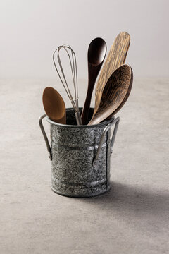 Rustic kitchen utensils including wooden spoons and a metal whisk in a vintage zinc pail on a gray stone surface, home cooking concept.