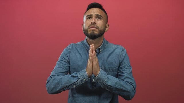 Young hispanic man with hands pressed in prayer gesture and closed eyes in a red studio, wearing denim shirt and beard; hope faith plea.