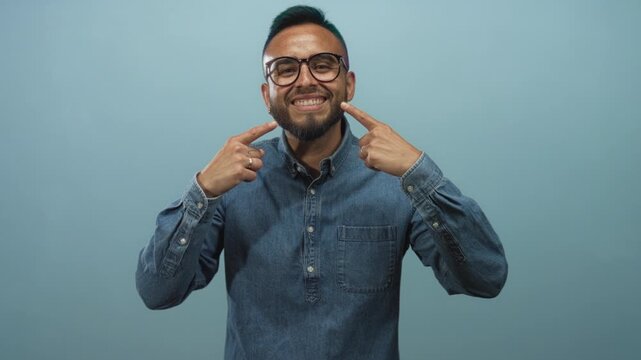 Man points fingers to his smile in blue studio while wearing glasses and a denim shirt, facing camera and showing teeth; joy confidence friendliness warmth.